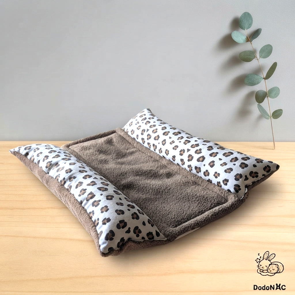 Lit pour petit animal DodoNac composé d'un matelas en sherpa marron et de deux traversins latéraux à motif léopard, posé sur une table en bois avec un fond gris uni. Une branche d'eucalyptus est visible en arrière-plan à droite, et le logo de la marque DodoNac avec deux lapins endormis est présent en bas à droite