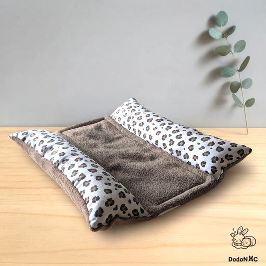 Lit pour petit animal DodoNac composé d'un matelas en sherpa marron et de deux traversins latéraux à motif léopard, posé sur une table en bois avec un fond gris uni. Une branche d'eucalyptus est visible en arrière-plan à droite, et le logo de la marque DodoNac avec deux lapins endormis est présent en bas à droite