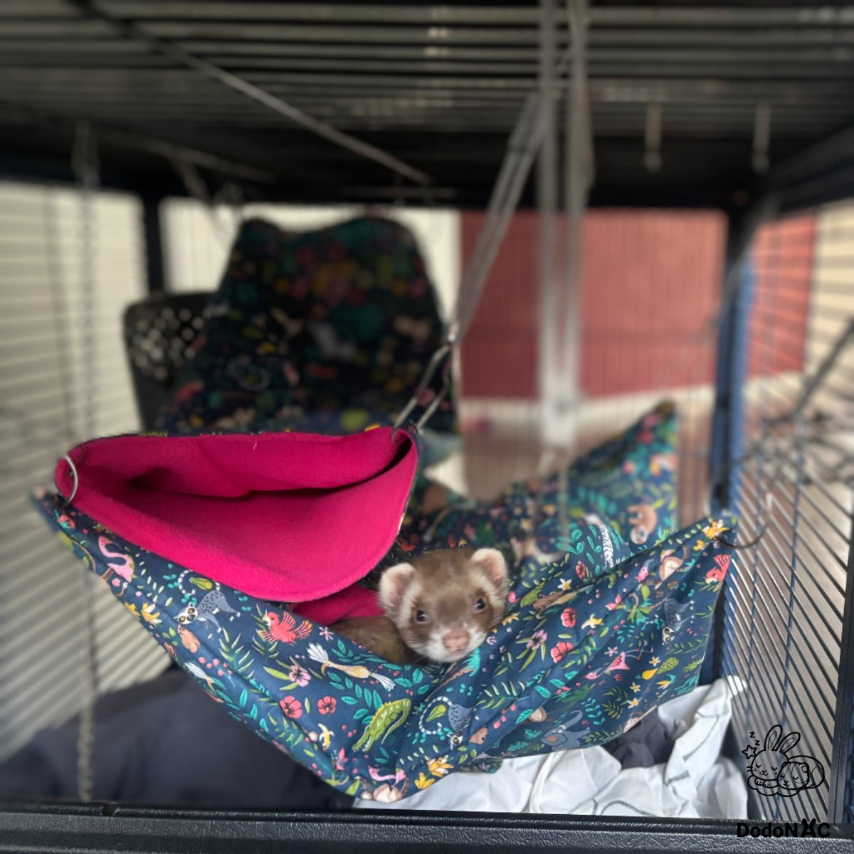 Furet brun blotti dans un hamac suspendu à motifs tropicaux et flamants roses à l'intérieur d'une cage métallique pour animaux de compagnie.