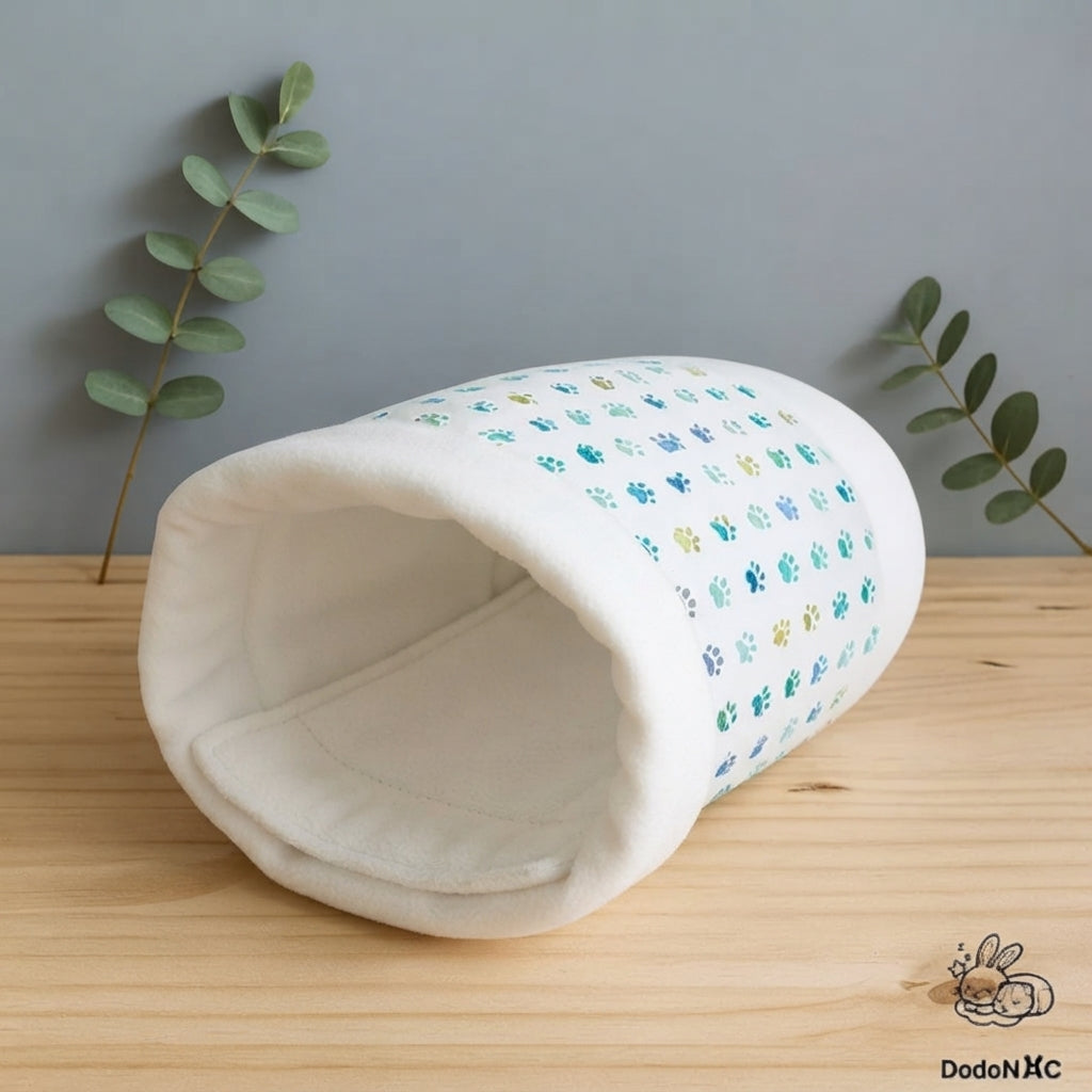 Tunnel cachette pour rongeurs et petits animaux fait main en tissu blanc à motifs de petites pattes bleues et vertes, posé sur un support en bois clair sur fond gris. Un pee pad en polaire blanche est installé à l'intérieur du tunnel.