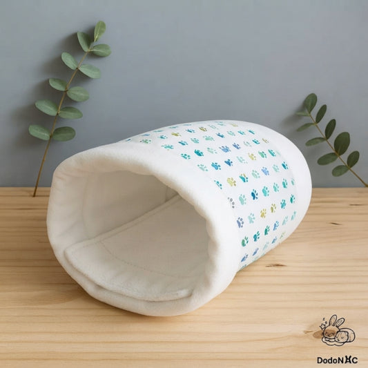 Tunnel cachette pour rongeurs et petits animaux fait main en tissu blanc à motifs de petites pattes bleues et vertes, posé sur un support en bois clair sur fond gris. Un pee pad en polaire blanche est installé à l'intérieur du tunnel.