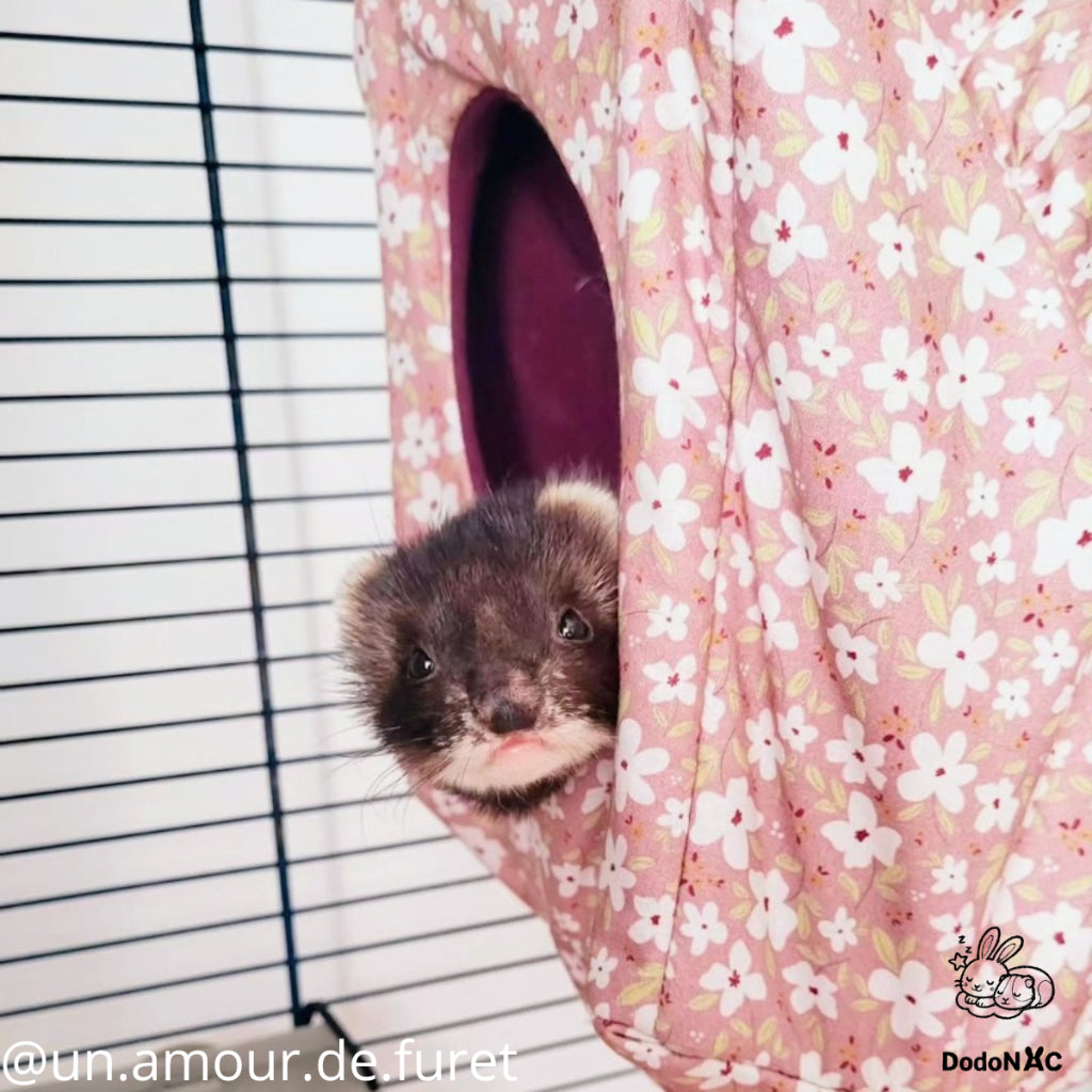 Un adorable furet brun et noir, avec le museau rose, regarde hors d'une cabane de couchage en tissu rose à motifs floraux blancs. L'accessoire est suspendu dans une cage à barreaux. Image créditée @un.amour.de.furet et DodoNAC
