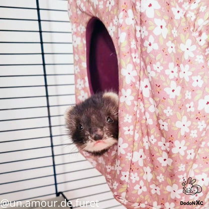 Un adorable furet brun et noir, avec le museau rose, regarde hors d'une cabane de couchage en tissu rose à motifs floraux blancs. L'accessoire est suspendu dans une cage à barreaux. Image créditée @un.amour.de.furet et DodoNAC